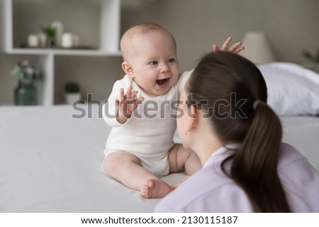Happy excited baby laughing at mom face, showing positive emotions, waving hands, sitting on bed, playing. New mother talking to charming infant child, cuddling kid, having fun Royalty-Free Stock Photo #2130115187