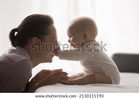 Joyful young mom playing with baby at home, enjoying motherhood, maternity leave. Cute infant child sitting on bed, touching happy mothers face, pushing nose, laughing Royalty-Free Stock Photo #2130115190