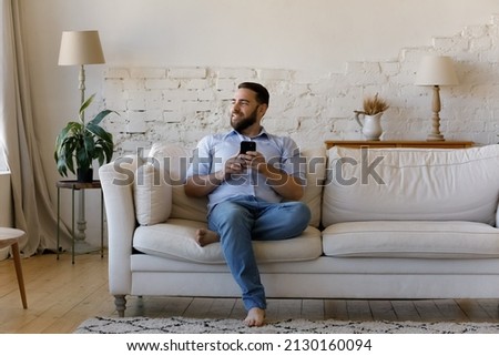 Smiling man sit on sofa at home with smartphone distracted from device look into distance spend weekend using modern cellphone, single guy enjoy e-dating services. Wireless tech, fun, leisure concept Royalty-Free Stock Photo #2130160094