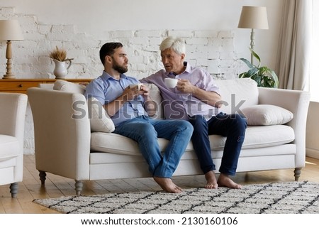 Serious older grey-haired man his grown up 35s sit on sofa holding cups drink tea or coffee talking, share news, communicating together on weekend at modern home. Care, family ties, support concept Royalty-Free Stock Photo #2130160106
