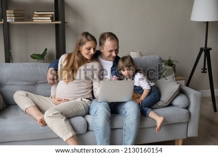 Happy expecting parents and little daughter kid relaxing on couch together, watching online movie on laptop, enjoying internet TV, making video call. Pregnant mom holding big tummy. Family leisure