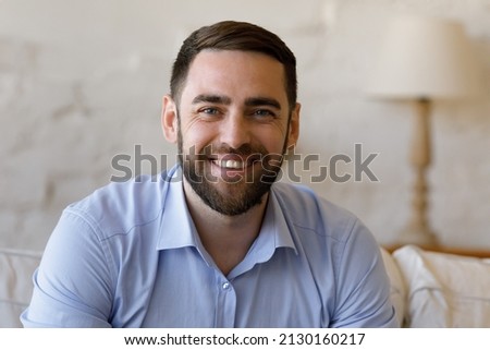 Head shot portrait millennial man sit on sofa at home smile look at camera, having attractive appearance, optimistic mood. Profile picture person take part in video conference event, e-dating services