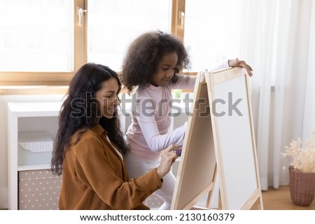 Happy bonding two female generations African American family, young mother and teenage kid daughter drawing with colored felt-tip pens on canvas easel or white board, enjoying hobby activity concept.