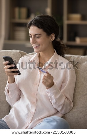 Vertical image joyful beautiful young Hispanic woman making payments in mobile shopping application using bank credit card, buying goods in internet store, enjoying purchasing services online.