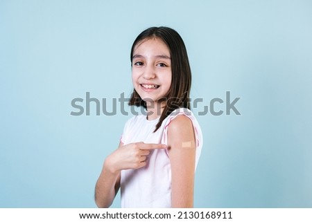 Hispanic girl child portrait after getting a vaccine protection and showing her arm with bandage receiving vaccination on blue background in Mexico Latin America Royalty-Free Stock Photo #2130168911
