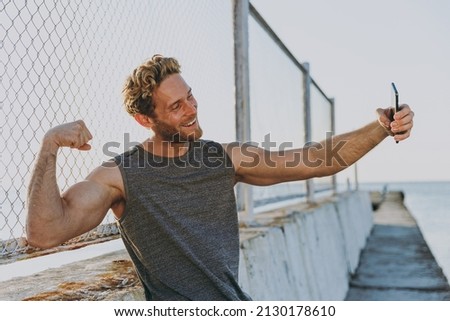 Young strong sporty athletic fit sportsman man in sports clothes warm up train do selfie shot mobile cell phone show muscles at sunrise sun over sea beach outdoor on pier seaside in summer day morning