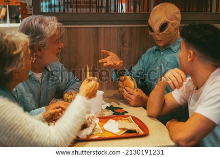 Happy family with ufo alien estraterrestrial guest enjoy together hamburgers in fast food. Concept of friendship and diverse people having fun together. Alien explaining at humans Royalty-Free Stock Photo #2130191231