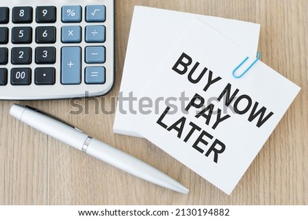 The words Buy Now Pay Later in text on a card on a wooden table next to a calculator, pen and note-taking paper