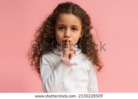 Portrait of adorable little girl making silence gesture with finger on lips, looking at the camera with smile. Indoor studio shot isolated on pink background 