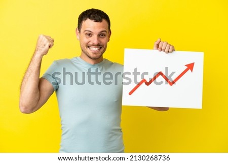 Brazilian man over isolated purple background holding a sign with a growing statistics arrow symbol and celebrating a victory
