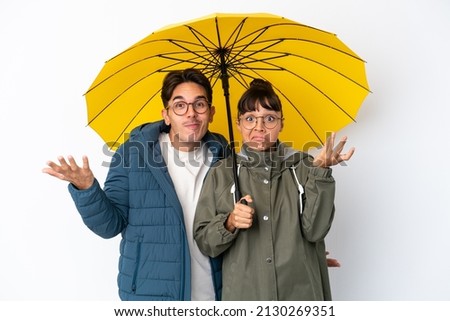 Young mixed race couple holding an umbrella isolated on white background having doubts while raising hands and shoulders Royalty-Free Stock Photo #2130269351