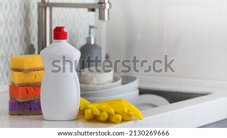 A white bottle with dishwashing gel, sponges and rubber gloves on the background of a sink with dirty dishes. Copy space Royalty-Free Stock Photo #2130269666