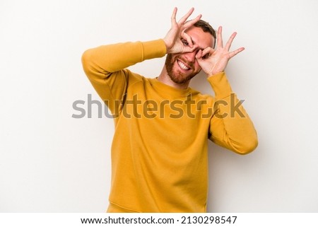 Young caucasian man isolated on white background showing okay sign over eyes