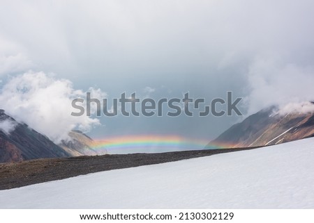 Minimalist alpine landscape with vivid rainbow. Scenic minimal view from snow hill to bright rainbow in mountain valley. Top view from snow mountain to colorful rainbow and low clouds in rainy weather