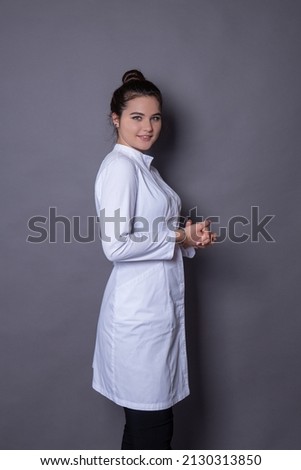 A young brunette girl with her hair taken in a bundle in a medical robe stands half a turn on a gray background folding her hands