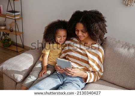 Two beautiful black girls of different age playing a game on the electronic device at home. Loving sisters sitting together on the couch streaming a show on a tablet. Background, close up, copy space.