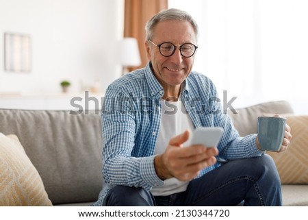 Portrait of smiling mature man in eyeglasses using mobile phone, watching video and holding cup of coffee, sitting on the couch in living room. Guy browsing internet, surfing web, free copy space Royalty-Free Stock Photo #2130344720