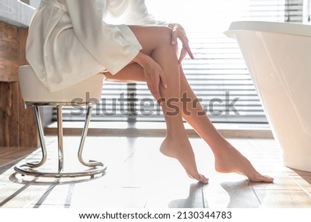 Body Hair Removal. Unrecognizable Woman Touching Smooth Skin On Legs After Depilation Treatment, Young Female In Silk Robe Sitting On Chair In Modern Bathroom At Home, Side View, Cropped Shot Royalty-Free Stock Photo #2130344783