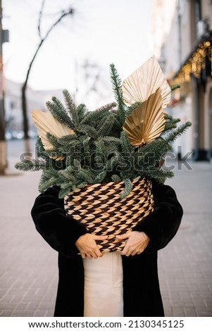 Woman standing in the street, holding big and beautiful bunch of Nobilis spruce wrapped in craft paper inside a wicker basket, vertical photo