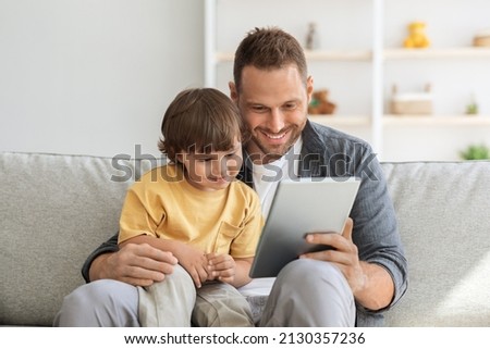 Child development content. Young positive father showing digital tablet to his cute little son, kid enjoying educational show online, sitting on sofa ta home