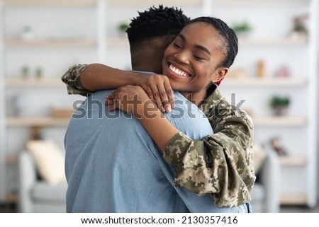 Closeup portrait of cheerful black female soldier lady in camouflage uniform hugging her boyfriend, home interior. Military wife came back from army, embracing her loving husband, finally together Royalty-Free Stock Photo #2130357416