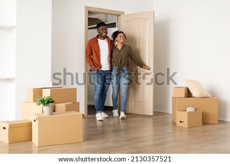 Real Estate Purchase. Happy African American Couple Entering Their New Home Standing Among Cardboard Moving Boxes In Empty Living Room Indoor. Housing For Young Family, Apartment Rent And Ownership Royalty-Free Stock Photo #2130357521