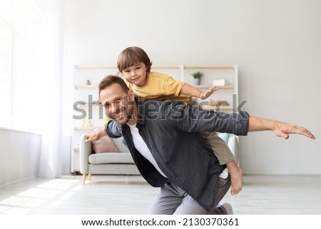 Games at home. Happy little boy riding daddy's back at home, kid playing plane together with father, smiling to camera at living room, free space Royalty-Free Stock Photo #2130370361