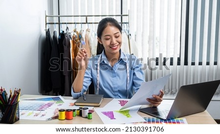 Asian woman fashion designer holding paper with clothes sketch of new collection dress and smiling while sitting in her workshop