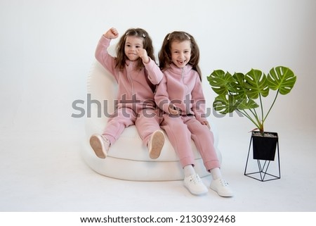 Two cute little girls in a pink overalls sits in a white chair. White background. Fashion. Childhood.