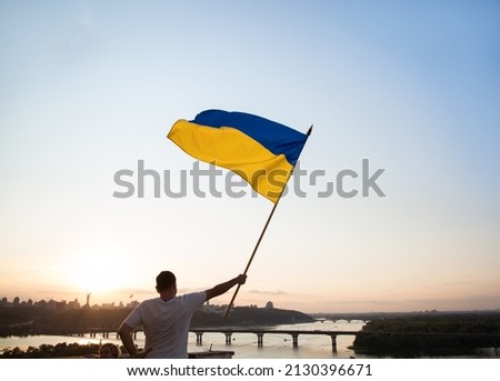 Ukrainian flag flutters in the wind against the sunset sky. A man standing on the roof of a house in Kyiv holds a large flag. National symbol of freedom and independence. Stop the war. Hope and Faith Royalty-Free Stock Photo #2130396671