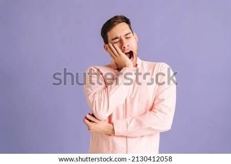 Portrait of exhausted and sleepy young handsome man yawning standing on purple isolated background in studio. Bored male feels sleepy, yawns as feels tired, opens mouth widely, closed eyes. Royalty-Free Stock Photo #2130412058