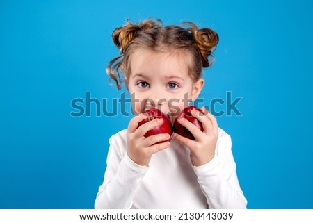 cute little girl with a funny hairstyle in a white T-shirt is holding two red apples in her hands. blue background. useful products for children. healthy snack. space for text. High quality photo