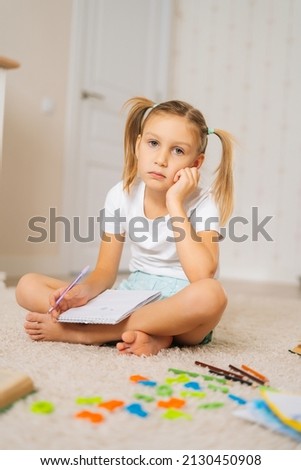 Vertical shot of tired primary little child girl writing homework in notebook sitting on floor at home looking at camera. Portrait of exhausted elementary schoolgirl taking notes in copybook.