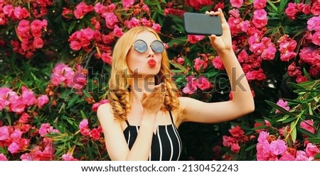 Beautiful woman taking selfie picture by phone blowing her red lips sending sweet air kiss on pink flowers roses background