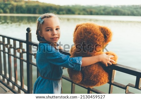 Portrait of girl embracing a cute teddy bear, holding her hands on a metal fence, looking over shoulder to camera smiling in denim clothes. Kids friendship concept. Happy kid. Copy space available.