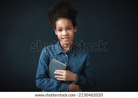 Happy pretty black teenage girl looking at camera with toothy smile with a book. Beautiful young curly haired schoolgirl.