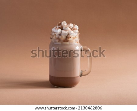 A cup of hot cocoa drink in clear glass with white marshmallows on a beige background. Milk drink.