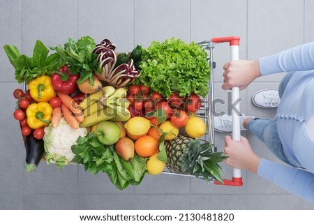 Woman pushing a shopping cart full of vegetables and fruits, healthy food and grocery shopping concept, top view Royalty-Free Stock Photo #2130481820
