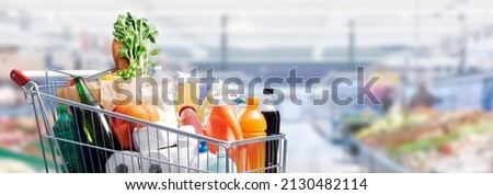 Shopping cart filled with food and drinks and supermarket shelves in the background, grocery shopping concept Royalty-Free Stock Photo #2130482114