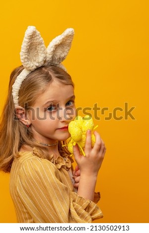 happy Easter. A cute little girl in a light dress with bunny ears holds painted eggs on a yellow background in her hands. The child smiles and puts on rabbit ears on Easter day.