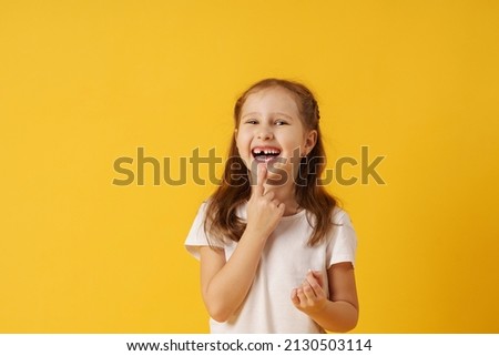 cute smiling preschool girl holds her first fallen baby tooth in her hand while standing on yellow background. growing permanent tooth is in the open mouth. . The concept of hygiene of baby teeth. Royalty-Free Stock Photo #2130503114