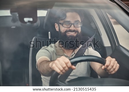 A Young attractive man driving a vehicle, looking at scenery, seen through the windshield glass. Happy driver, holding hands on the steering wheel with a wide beautiful smile. Royalty-Free Stock Photo #2130515435