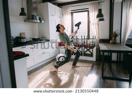 Young happy man doing cleaning and dancing with vacuum cleaner in kitchen and living room at home. A handsome guy is vacuuming a carpet, dancing and singing in a cozy room on a sunny day Royalty-Free Stock Photo #2130550448