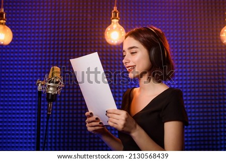 Young woman with headphones is reading from paper and recording a podcast in an audio recording studio Background from acoustic panel, blue light and vintage light bulbs.