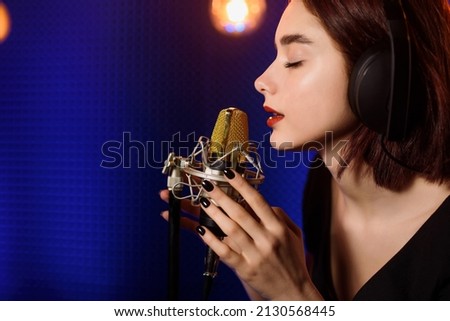 Young woman with headphones is recording a song in an audio recording studio. Background from acoustic panel, blue light and vintage light bulbs. Copy space for your text. 