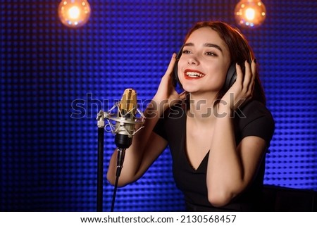 Young happy smiling woman singer with headphones  is recording a song in an audio recording studio. Background from acoustic panel, blue light and vintage light bulbs. Copy space for your text. 
