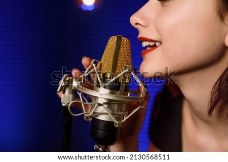 Young woman singer  is recording a song in an audio recording studio. Background from acoustic panel, blue light and vintage light bulbs. Copy space for your text. 
