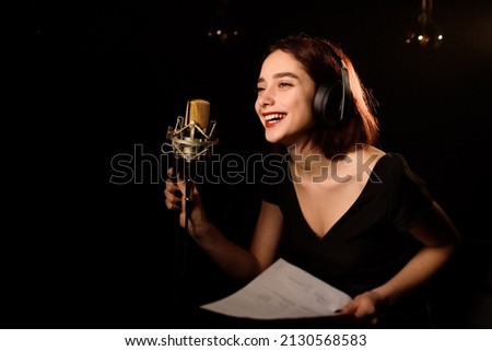 Young woman with headphones is reading from paper and recording a podcast in an audio recording studio on black bakcground. Copy space for your text