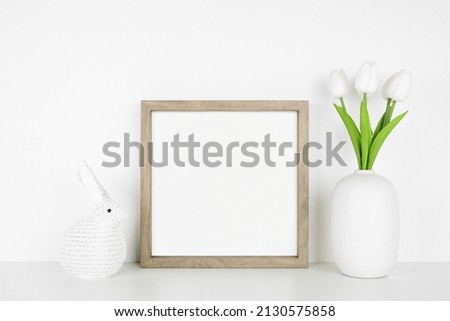 Mock up wood frame with Easter decor on a white shelf. Knit bunny and tulip flowers. Square frame against a white wall. Copy space.