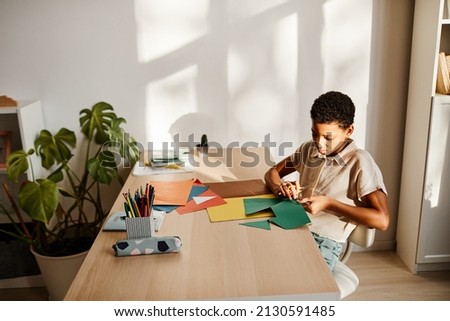 Minimal portrait of young black girl crafting with colored paper at desk lit by sunlight, copy space